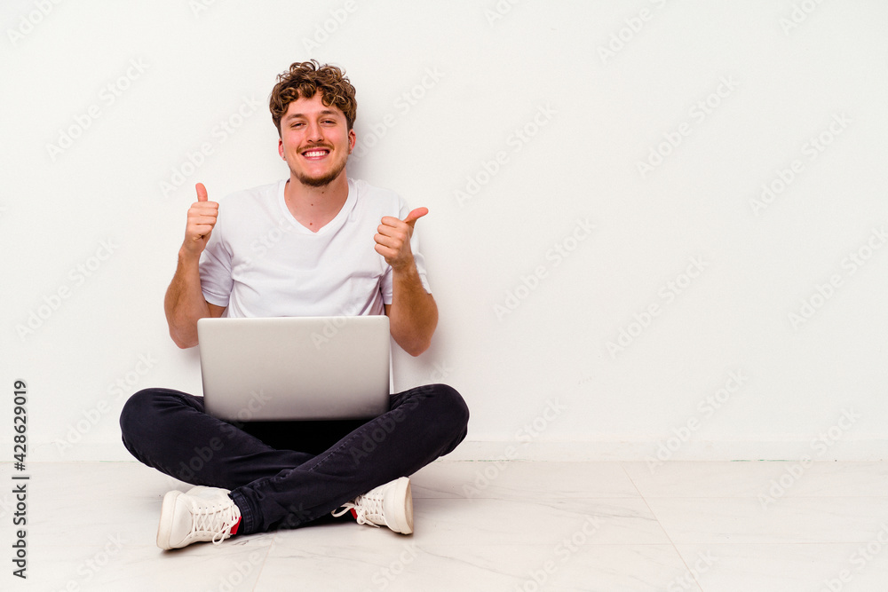 Young caucasian man sitting on the floor holding on laptop isolated on white background raising both thumbs up, smiling and confident. Young caucasian man sitting on the floor holding on laptop isolated on white background raising both thumbs up, smiling and confident.