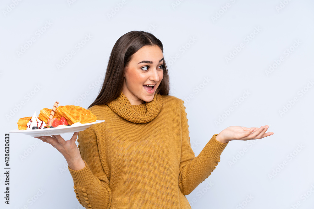 Young caucasian woman holding waffles isolated on blue background with surprise facial expression Young caucasian woman holding waffles isolated on blue background with surprise facial expression