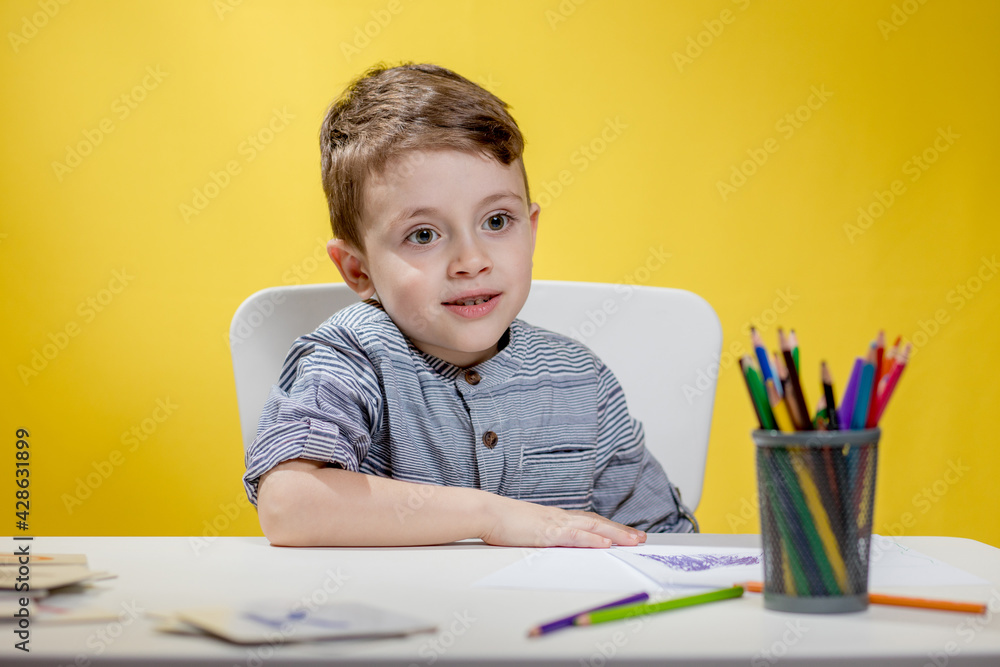 Smiling little boy at the table draw with crayons for mum on yellow background. Creativity concept. Back to school. Smiling little boy at the table draw with crayons for mum on yellow background. Creativity concept. Back to school.