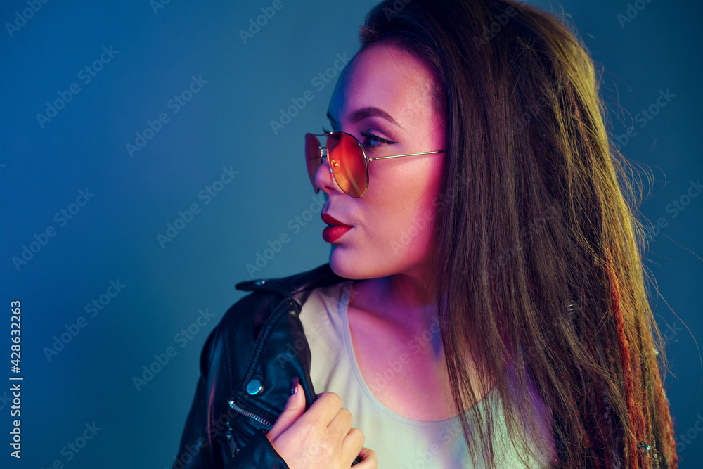 Fototapeta premium Asian girl in sunglasses and black leather jacket posing against dark blue background