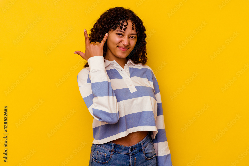 Young mixed race woman isolated on yellow background showing rock gesture with fingers Young mixed race woman isolated on yellow background showing rock gesture with fingers