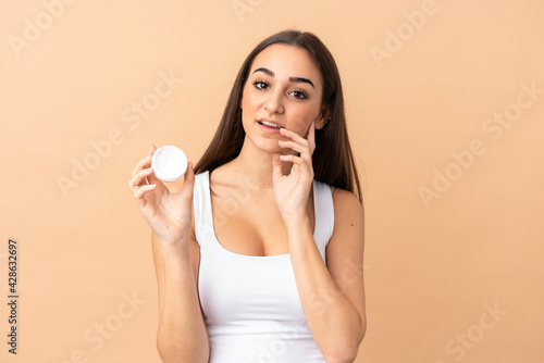 Young caucasian woman isolated on beige background with moisturizer