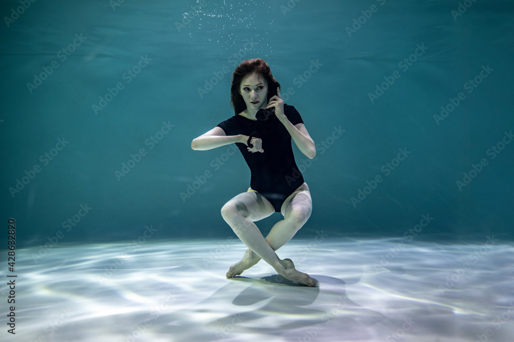 beautiful girl in a black swimsuit posing underwater with a black telephone receiver on a blue background 