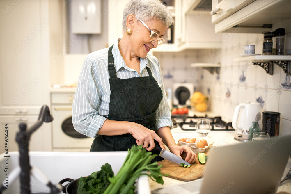 Technology, cookery and food concept. Happy senior woman in glasses and apron standing at kitchen counter, cutting cucumber for healthy meal, watching video recipe or favorite series online on laptop Technology, cookery and food concept. Happy senior woman in glasses and apron standing at kitchen counter, cutting cucumber for healthy meal, watching video recipe or favorite series online on laptop