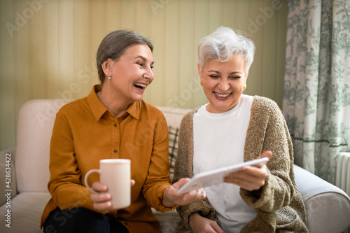 Two stylish mature female friends enjoying wireless high-speed internet connection, using tablet computer, looking through old digital pictures and laughing, being in good mood, having over coffee photo