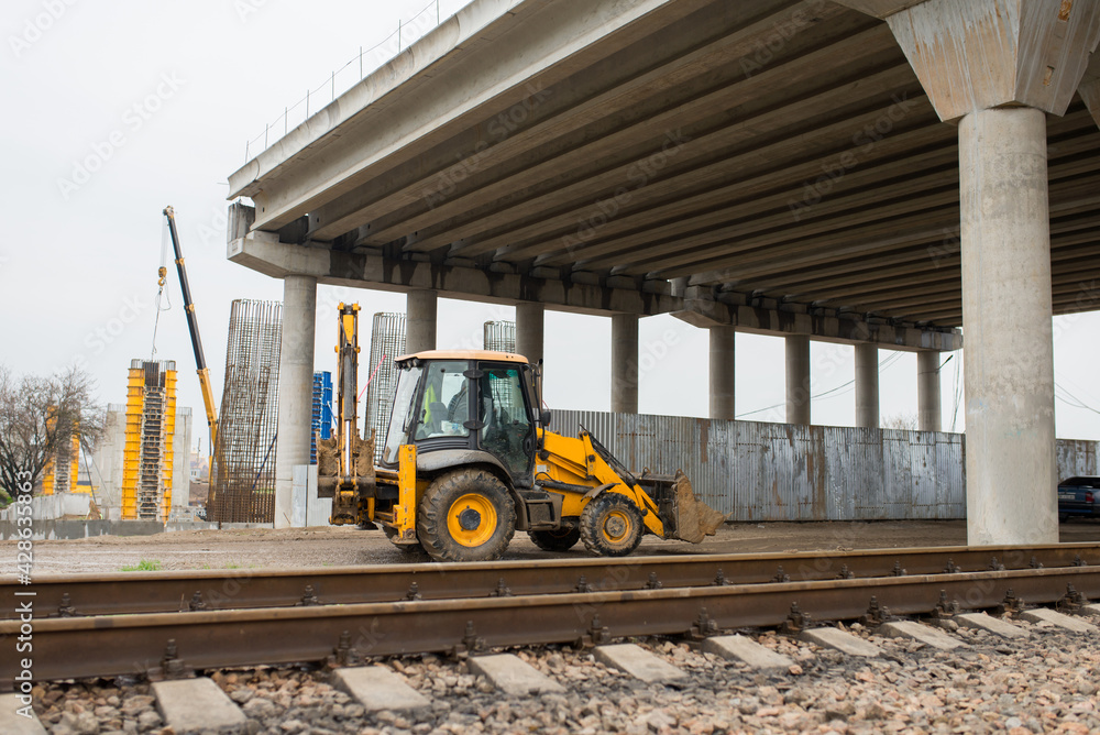 Obraz premium A construction excavator passes under a reinforced concrete road bridge under construction