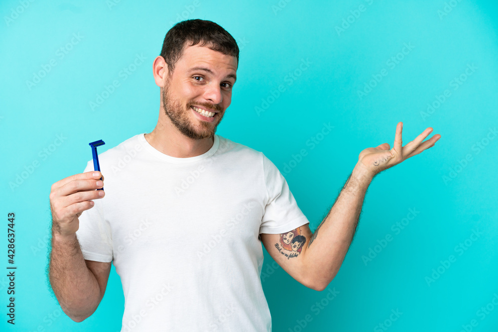Brazilian man shaving his beard isolated on blue background extending hands to the side for inviting to come Brazilian man shaving his beard isolated on blue background extending hands to the side for inviting to come