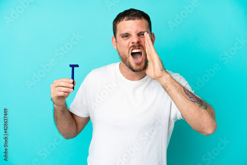 Brazilian man shaving his beard isolated on blue background shouting with mouth wide open