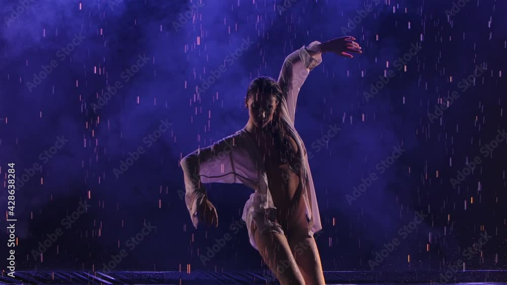 Contemporary dance ballerina dances sensually in rain on water surface, creating splashes. Silhouette of wet woman in white shirt move plastically in dark studio with blue light. Slow motion. Close up Contemporary dance ballerina dances sensually in rain on water surface, creating splashes. Silhouette of wet woman in white shirt move plastically in dark studio with blue light. Slow motion. Close up