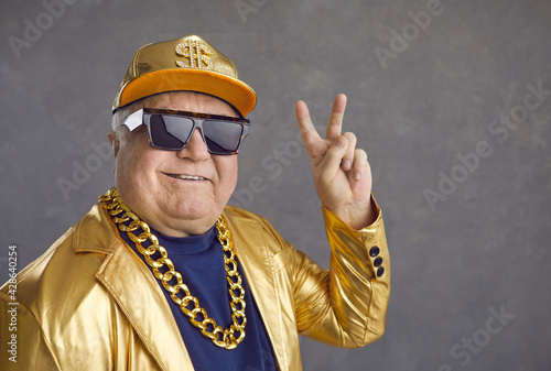 Grandpa is trying to be young. Funny and cheerful senior man in luxury clothes showing v-sign on gray background. Man in sunglasses is wearing a gold jacket and cap and wears a chain around his neck.