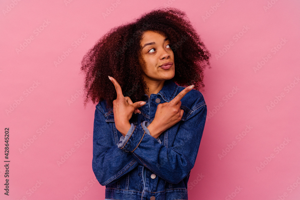 Young african american woman isolated on pink background points sideways, is trying to choose between two options. Young african american woman isolated on pink background points sideways, is trying to choose between two options.