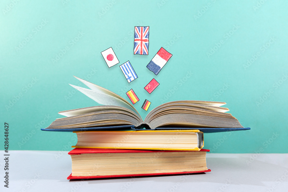 On the table is a stack of books, with an open book on top, from which flags fly out. Turquoise background. The concept of learning foreign languages - obrazy, fototapety, plakaty 
