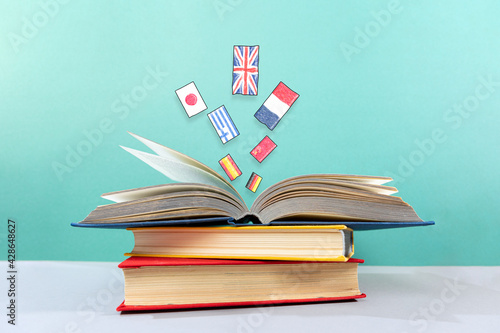 On the table is a stack of books, with an open book on top, from which flags fly out. Turquoise background. The concept of learning foreign languages
