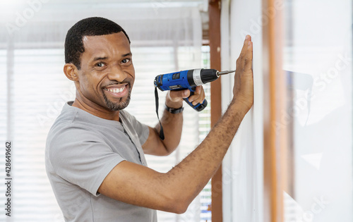 Portrait of happy African American black man using electric drill on wall. DIY, do it yourself hobby, handyman worker using drill while working on construction site. Home interior builder moving 