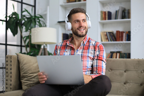 Concentrated young freelancer businessman sitting on sofa with laptop, working remotely online at home.