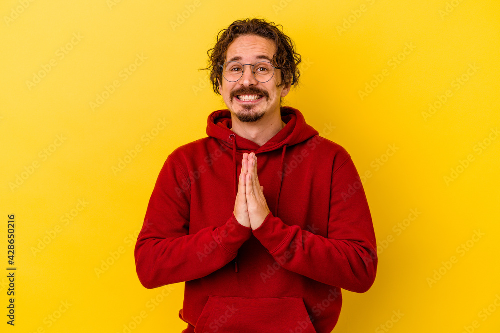 Fototapeta premium Young caucasian man isolated on yellow background holding hands in pray near mouth, feels confident.