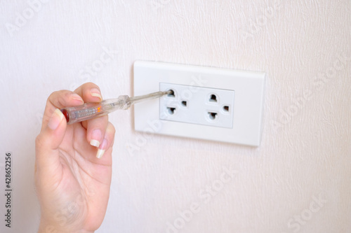 An electrician is hand holding on voltage electrical tester screwdriver with a red LED lamp into a hole of an electric socket to determine