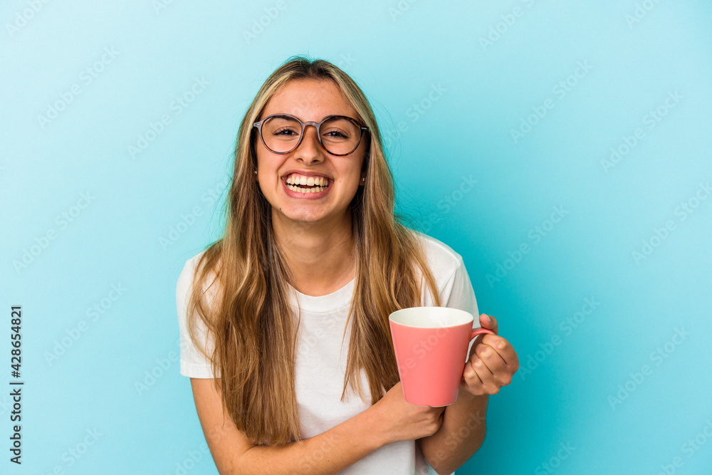 Young caucasian blonde woman holding a mug isolated on blue background laughing and having fun. Young caucasian blonde woman holding a mug isolated on blue background laughing and having fun.