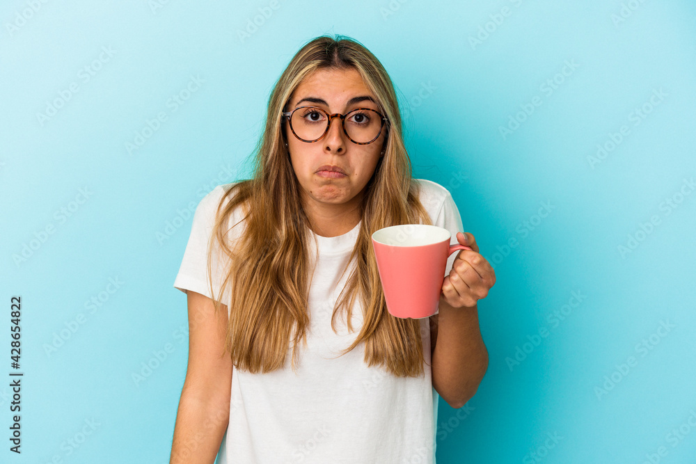 Young caucasian blonde woman holding a mug isolated on blue background shrugs shoulders and open eyes confused. Young caucasian blonde woman holding a mug isolated on blue background shrugs shoulders and open eyes confused.
