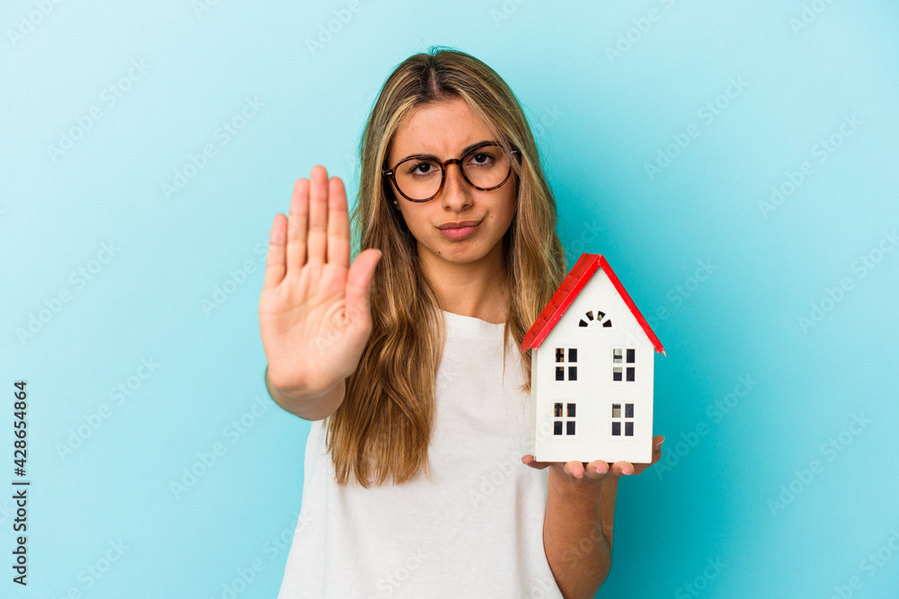 Young caucasian woman holding a house model isolated on blue background standing with outstretched hand showing stop sign, preventing you. Young caucasian woman holding a house model isolated on blue background standing with outstretched hand showing stop sign, preventing you.