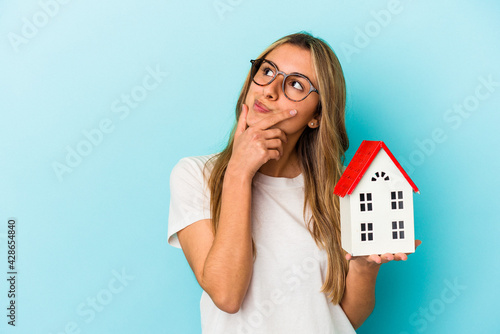 Fotografija Young caucasian woman holding a house model isolated on blue background looking sideways with doubtful and skeptical expression