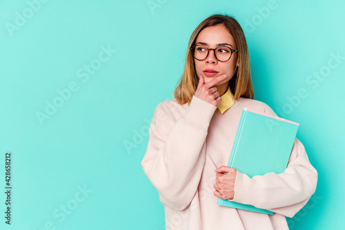Young student woman holding books isolated on blue background looking sideways with doubtful and skeptical expression.