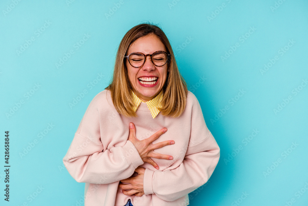 Young student caucasian woman isolated on blue background laughs happily and has fun keeping hands on stomach. Young student caucasian woman isolated on blue background laughs happily and has fun keeping hands on stomach.