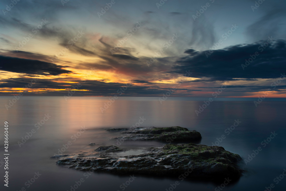 Cloudy sunset reflecting into an calm dark ocean with boulders in the foreground at island of Gotland in Sweden