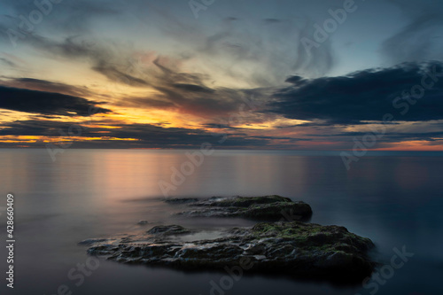 Cloudy sunset reflecting into an calm dark ocean with boulders in the foreground at island of Gotland in Sweden
