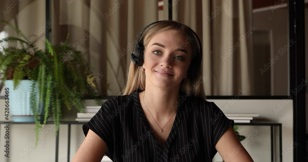 Web camera view smiling young beautiful blonde business lady in wireless headset with microphone looking at camera, holding video call zoom meeting, negotiating working issues with colleagues online. Web camera view smiling young beautiful blonde business lady in wireless headset with microphone looking at camera, holding video call zoom meeting, negotiating working issues with colleagues online.
