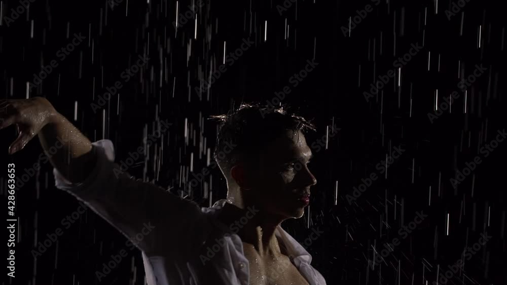 Silhouette of wet male dancer performing modern ballet dance in rain and splashing water. Male sensual modern choreography on black background in center of light beam. Slow motion. Close up. Silhouette of wet male dancer performing modern ballet dance in rain and splashing water. Male sensual modern choreography on black background in center of light beam. Slow motion. Close up.