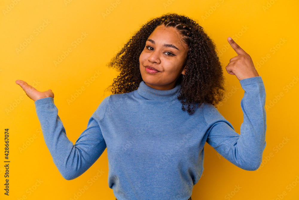 Young african american woman isolated on yellow background holding and showing a product on hand. Young african american woman isolated on yellow background holding and showing a product on hand.