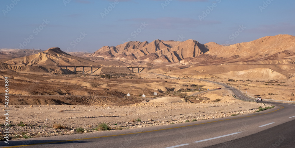Asphalt road going through the Negev Desert near Big Crater in Israel. Chain of varicolored mountain ridges on the background. Dramatic, colorful and diverse desert landscape. Bridge above the road.