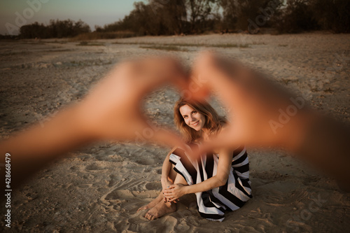 Young woman sits on the beach in the rays of sunset and smile. Child shows a smiling mother a heart by hands gesture, symbol of love. Relationship parents and children. Mother day. Family confidence