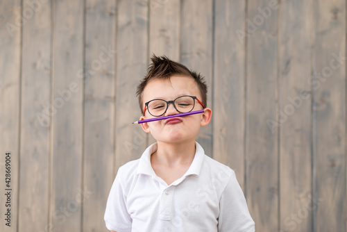 Portrait of little boy wearing glasses and white T-shirt at wooden background. child holding a pencil with his lips. Preparation or back to school.