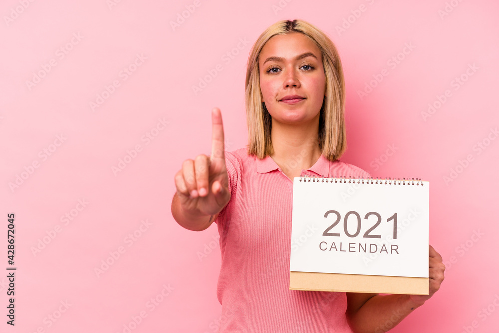 Young venezuelan woman holding a calendar isolated on pink background showing number one with finger. Young venezuelan woman holding a calendar isolated on pink background showing number one with finger.