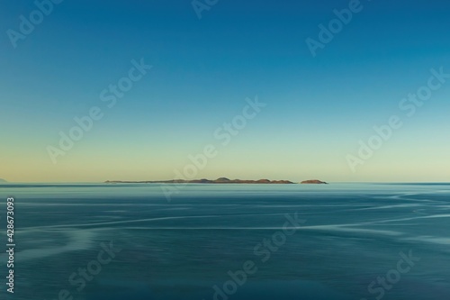 Long exposure photograph, blue water blue sky, small island in the distance, minimalism.
