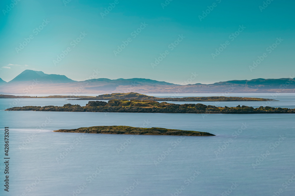 Long exposure photograph, blue water, blue sky, three small islands, mountain range in the distance.