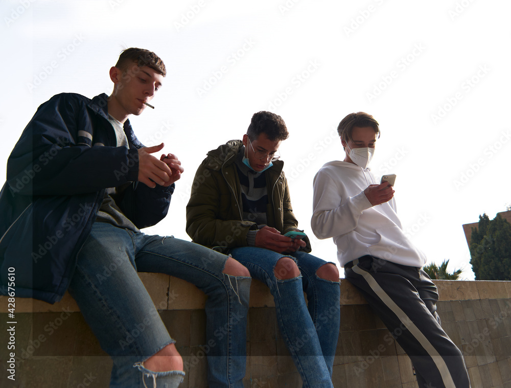 A closeup of three young Caucasian and Hispanic guys sitting outdoors smoking and using their phones wearing sanitary masks A closeup of three young Caucasian and Hispanic guys sitting outdoors smoking and using their phones wearing sanitary masks