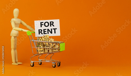 House for rent symbol. Miniature shopping cart with wooden houses, words for rent. Wooden model of human. Beautiful orange background, copy space. Business and house for rent concept.