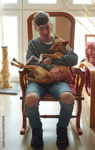 A vertical shot of a young Caucasian guy sitting hugging an adorable Pharaoh Hound