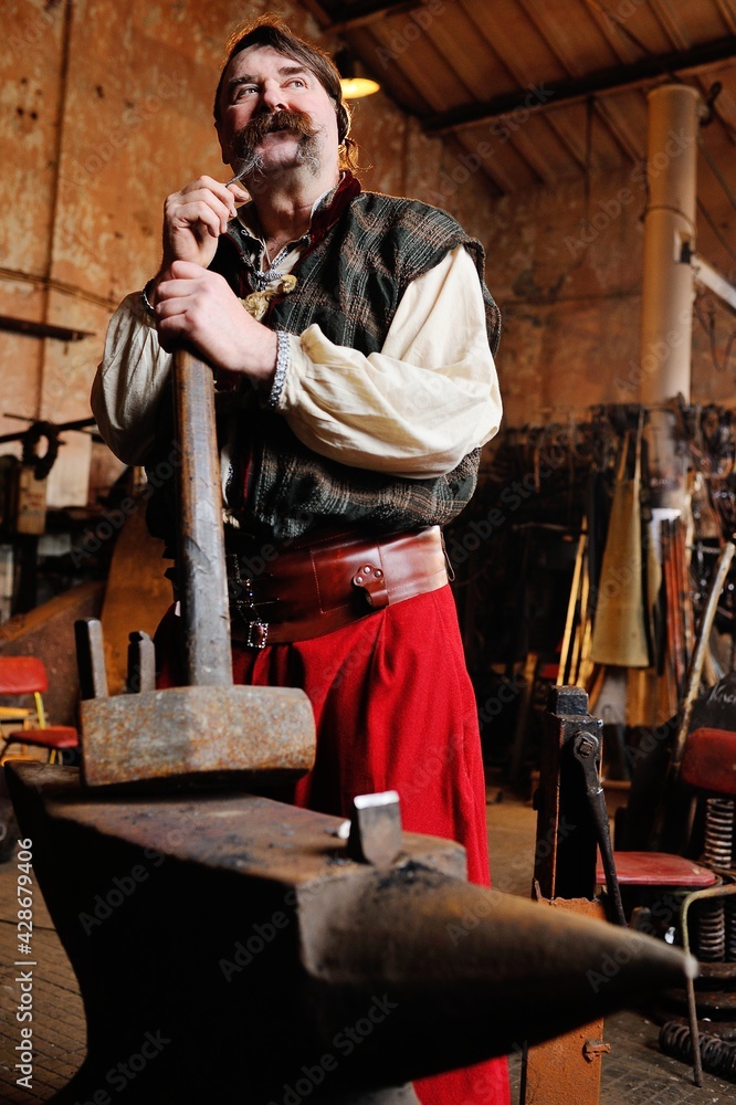 portrait of a blacksmith with a beautiful thick mustache with a huge hammer in the hands on the background of the forge.