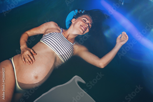 Foto Beautiful pregnant woman floating in tank filled with dense salt water used in meditation, therapy, and alternative medicine