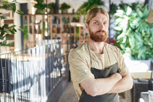 Waist up portrait of bearded cafe worker wearing apron and looking at camera while standing with arms crossed in eco-friendly green interior, copy space