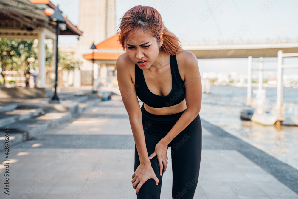 Young Asian female athlete runner in sportswear at river side suffering pain and discomfort from knee injury during morning run