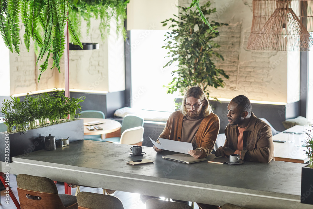 © Seventyfour - Wide angle portrait of two contemporary men collaborating on project during business meeting in green office or cafe interior, copy space