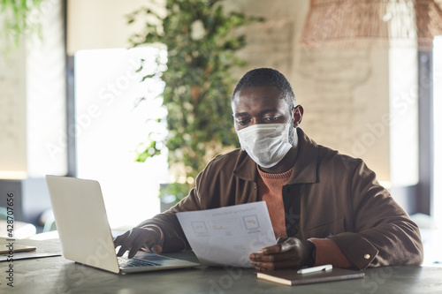 Portrait of African-American man wearing mask while using laptop and working in cafe or office, copy space