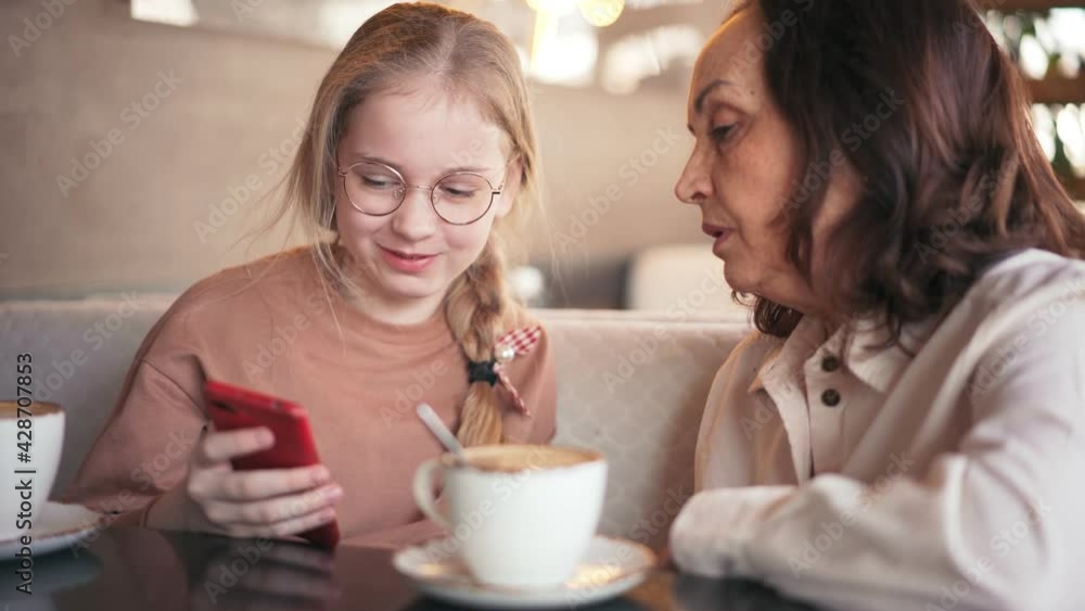 Happy grandmother and cute granddaughter using mobile phone together, while sitting at the cafe. Smiling senior grandmother and little girl having fun spending time together. Happy grandmother and cute granddaughter using mobile phone together, while sitting at the cafe. Smiling senior grandmother and little girl having fun spending time together.