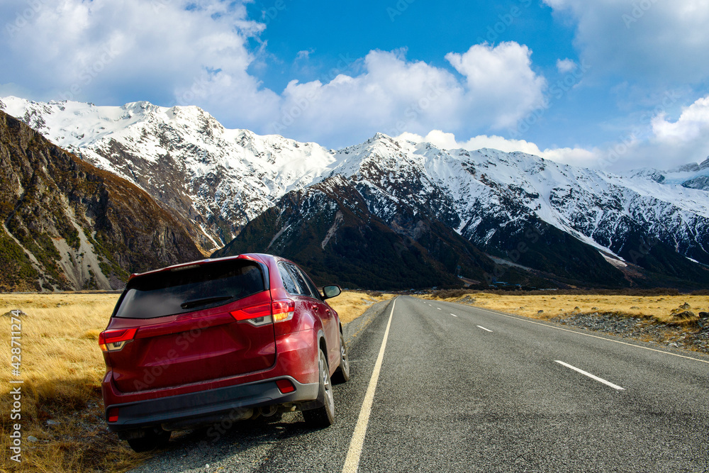 Red SUV car on the roadside, background is big and tall Mountains in winter, dry grass turns yellow in blue skies and clouds in the daytime in Mount cook National Park in southern island, New Zealand Red SUV car on the roadside, background is big and tall Mountains in winter, dry grass turns yellow in blue skies and clouds in the daytime in Mount cook National Park in southern island, New Zealand