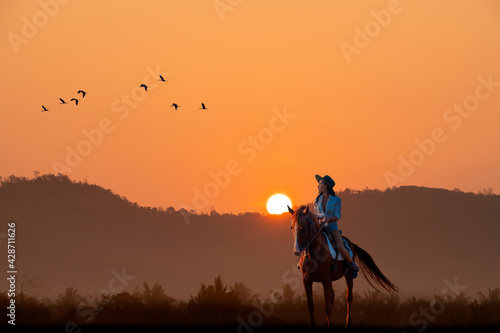 Cowgirl rider on horse back with silhouette mountain with flock of birds flying with sun sky background.